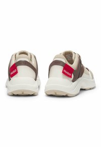 Beige and brown sneakers with a textured upper, padded collar, and contrasting red logo detail on the heel. Chunky white sole.
