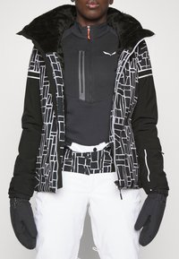 Veste de ski noire isolée avec un motif géométrique blanc, col doublé de fourrure et poche avant zippée. Associée à un pantalon de ski blanc et des moufles noires.
