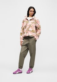 Γυναίκα που φοράει ροζ, πράσινο και λευκό φλοράλ φλις μπλουζάκι, χακί παντελόνι με κορδόνι, μοβ αθλητικά παπούτσια και γυαλιά με χρωματιστά φακούς, στέκεται μπροστά σε λευκό φόντο.