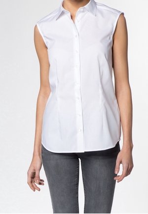 Vrouw draagt een mouwloos wit overhemd met knoopsluiting, gecombineerd met donkergrijze nauwsluitende spijkerbroek, staand tegen een effen achtergrond.