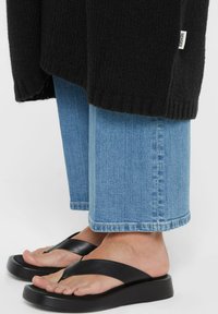 Person, der eine verkürzte blaue Jeans, schwarze Flip-Flops und einen schwarzen gestrickten Pullover mit einem weißen "SENSES"-Label trägt.