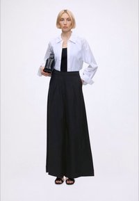 Femme aux cheveux blonds courts portant une chemise blanche, un pantalon noir à jambes larges, un top noir, tenant une pochette noire, debout sur fond blanc.
