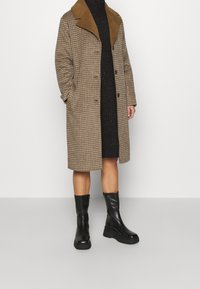 Cappotto a scacchi marrone con colletto beige, indossato sopra un abito di maglia nero. Abbinato a stivaletti neri in pelle, con una robusta piattaforma.
