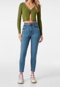 Olivgrön ribbade kort tröja med V-ringning och framknäppning, kombinerad med ljusblå högmidjade jeans med lätt slitningar.