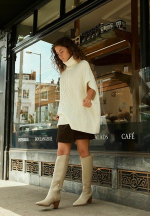 Poncho bianco oversize lavorato a maglia con motivo a treccia, abbinato a una gonna marrone e stivali in cuoio beige al ginocchio, ambientato in un caffè.