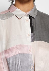 Gros plan sur une personne portant une chemise boutonnée à col pointu, avec un motif géométrique aux tons rose, noir et beige.
