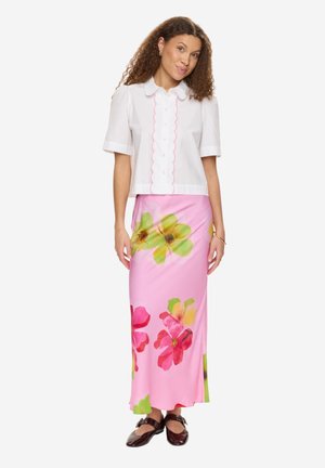 Kobieta o kręconych włosach, ubrana w biały krótki rękaw koszuli z guzikami oraz różową długą spódnicę w zielone i czerwone kwiatowe wzory, stojąca z rozluźnionymi rękami.