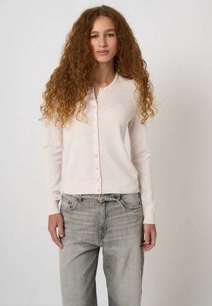 Kobieta z długimi kręconymi włosami, ubrana w bladoróżowy kardigan zapinany na guziki oraz jasnoszare dżinsy, stojąca na tle jednolitego jasnego tła.