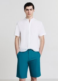 Chemise blanche à manches courtes avec col montant, associée à un short bleu sarcelle mi-genoux. Les deux ont une texture lisse et une coupe moderne.