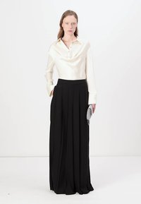 Camicetta in satin crema con scollo a cowl e bottoni frontali, abbinata a pantaloni neri plissettati a vita alta. Dettaglio aggiuntivo con pochette argento.