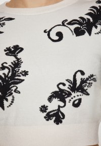 Pulover tricotat alb cu modele florale și cu răsuciri negre. Dispune de un gât rotund și manșete cu nervuri, fabricat dintr-un material moale.
