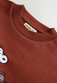 Brązowa bluza z ribbowym dekoltem, zdobiona białą grafiką rysunkowego psa oraz metką "PEANUTS" na wewnętrznej stronie kołnierza. Miękka tekstura materiału.