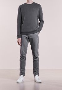 Sudadera gris con cuello redondo y mangas ajustadas, combinada con pantalones gris slim-fit y zapatillas blancas, de pie sobre un suelo claro.
