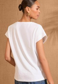 White cotton t-shirt with short sleeves and a relaxed fit, featuring a smooth texture and curved hem. Model wears denim jeans.