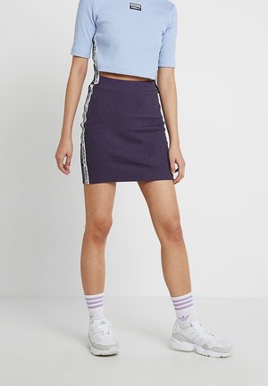 Femme portant une jupe violette ajustée et un haut court bleu clair avec les bandes du logo Adidas, des chaussettes blanches et des baskets blanches, debout devant un fond uni.