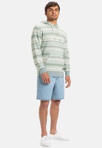 Sudadera con capucha a rayas verde claro con cordones, que presenta un bolsillo delantero. Combinada con pantalones cortos de mezclilla azul claro y zapatillas blancas.