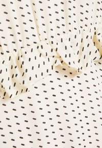 Světle béžová tkanina s černými tečkovanými vzory, s volánkovým lemem. Materiál se vyznačuje měkkou texturou a splývavým designem.