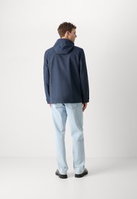 Navy waterproof jacket with a hood, featuring a smooth texture and a slightly loose fit, paired with light blue jeans and black shoes.
