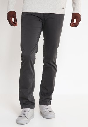 Mann trägt schwarze Slim-Fit-Hose, weiße Sneakers und ein hellgraues Langarmshirt, steht vor einem einfarbigen Hintergrund.