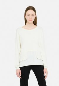 Maglione giallo chiaro a maniche lunghe con pannello frontale testurizzato, scollatura rotonda e due tasche frontali. Abbinato a pantaloni neri aderenti.