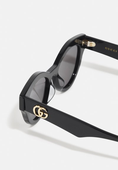 Sorte solbriller med rektangulære rammer, grå linser og guld "GG" logo accentuer på stængerne. Glat, blank plastmateriale.