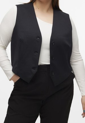 Vrouw draagt een zwart gilet met knopen over een witte top met lange mouwen en zwarte, hooggetailleerde broek, met één hand in de zak.