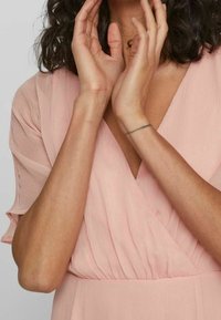 Žena oblečená v světlerůžových šatech s krátkými rukávy, s rukama zvednutýma blízko obličeje a částečně viditelnými kudrnatými tmavými vlasy.