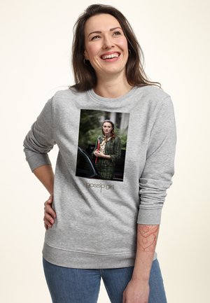 Femme souriante aux longs cheveux bruns portant un sweat-shirt gris avec une photo d'une femme en tenue verte tenant un dossier rouge, jean et tatouage.