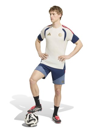 Ung mand i hvidt og blåt fodboldtøj står med den ene fod på en sort-hvid fodbold mod en hvid baggrund.