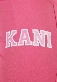 Rožinė raukšlėta medžiaga su žodžiu "KANI", atspausdintu ryškiomis baltomis raidėmis. Tekstas turi šiek tiek gradientinį efektą, kuris pagerina vizualinį patrauklumą.