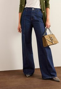 Jean large bleu foncé avec des coutures contrastantes et taille haute, associé à un sac à main texturé doré et des chaussures à bouts pointus.