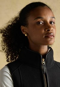 Čierna fleecová vesta s vysokým golierom a zlatým zipsom. Modelka má kučeravé vlasy a nosí malé zlaté náušnice. Neutrálny pozadí.