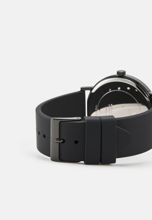 Brăţară de ceas din silicon negru cu un finisaj mat, dotată cu o cataramă metalică şi un cadran rotund vizibil din spate, având elemente de design minimalist.