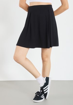 Vrouw draagt een zwarte geplooide rok, witte sokken en zwart-witte Adidas sneakers, staand met één been over het andere geslagen.