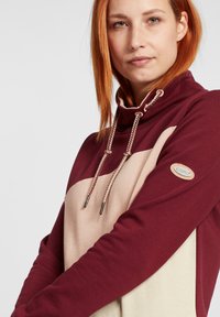 Burgunderfarget og beige hettegenser med snor, blandet stoff, med en logo-patch på ermet og fargeblokkdesign.