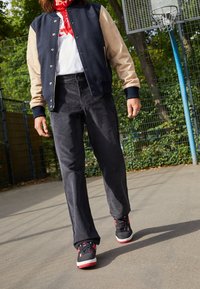 Czarne spodnie cord woskowe, granatowa i beżowa kurtka bomberka, biała koszulka, czerwona szalik, czarne sneakersy z czerwonymi akcentami, stojący na chodniku.