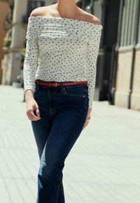 Top bianco scollato con spalle scoperte e pois neri, slim fit, abbinato a jeans a vita alta blu scuro e una cintura rossa. Ambientazione urbana informale.