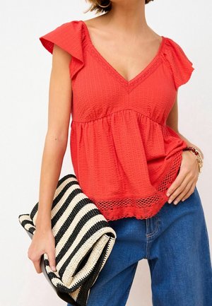 Femme portant un chemisier rouge texturé avec un bord en dentelle et un jean bleu, tenant une grande pochette tissée noire et blanche à rayures.