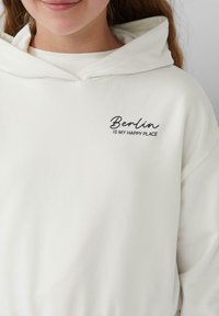 Weißer Hoodie aus weichem Stoff mit einer Fronttasche und einer Kordelzugkapuze. Bestickter Text: "Berlin IST MEIN GLÜCKSORT" in Schwarz.