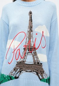 Helles blaues Sweatshirt mit gestricktem Eiffelturm, weißen Wolken, grünem Gras und rotem geschwungenem "Paris"-Text auf der Vorderseite.