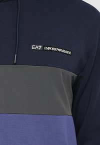 Sweatshirt com capuz em tons de azul-marinho, cinza e roxo. O tecido é suave, com um emblema próximo ao decote e cordão ajustável.