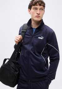 Ναυτικό μπλε τζάκετ φόρμας PUMA με λευκές ρίγες, εμπρός φερμουάρ και ψηλό γιακά, συνδυασμένο με μαύρη υφασμάτινη τσάντα duffel.