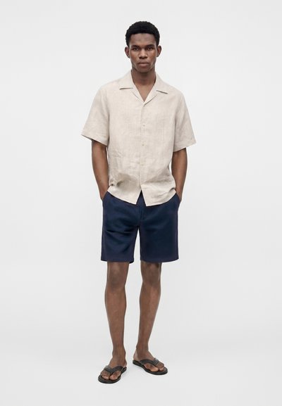 Homme portant une chemise beige à manches courtes boutonnée, un short bleu marine et des tongs noires, debout les mains dans les poches sur fond blanc.