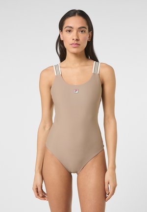 Kvinde iført en beige badedragt med stribede skulderstropper og et lille logo på brystet mod en ensfarvet baggrund.