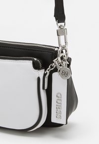 Sorte og hvide læderhåndtaske med lynlås lukning, sølvfarvet hardware, fremtrædende mærkeetiket og glat tekstur. Har et kontrasterende kantdesign.