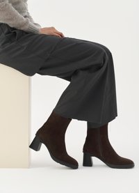 Brown suede ankle boots with a block heel and side zipper, paired with loose gray pants, sitting against a light-colored pedestal.