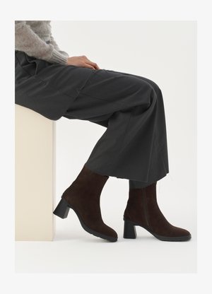 Hnedé semišové kotníkové čižmy s blokovým podpätkom a bočným zipsom, kombinované so voľnými sivými nohavicami, posadené pri svetlej podstave.