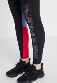 Zwarte sportleggings met meerkleurige zijpanelen (rood, blauw) en reflecterende branding. Gecombineerd met zwarte sportschoenen.