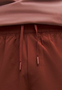 Prim-plan al unei perechi de pantaloni de culoare ruginie, cu talie elastică și șnururi asortate, purtați cu un top roz prăfuit, introdus în pantaloni.