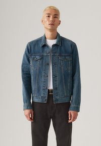 Donkerblauwe denimjack, met een klassiek ontwerp met borstzakken, metalen knopen en een licht vervaagde textuur. Gecombineerd met een wit T-shirt.